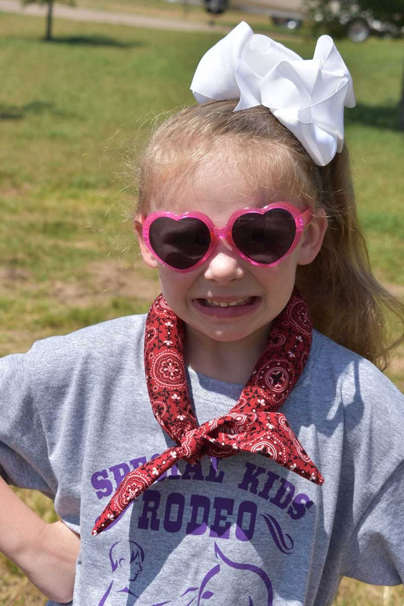 Young girl in pink heart sunglasses and white hair bow wearing a red bandana neck wrap, Special Kids Rodeo shirt